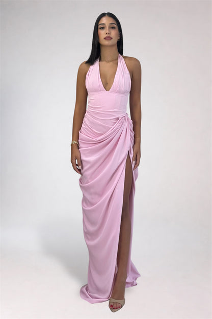 A woman modeling a light pink halter-neck maxi dress with a draped wrap-style skirt and a high front slit, photographed against a plain light gray studio background.