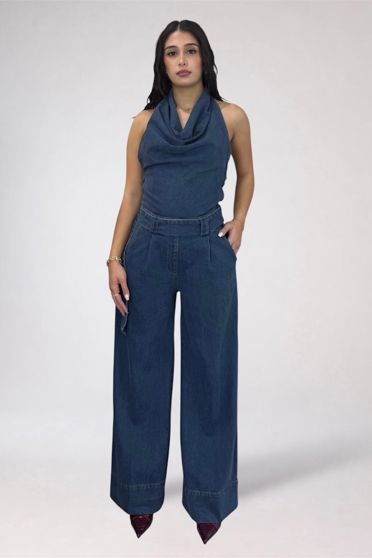 A woman modeling a dark blue denim cowl-neck sleeveless top paired with matching wide-leg denim pants, standing against a light gray studio background.