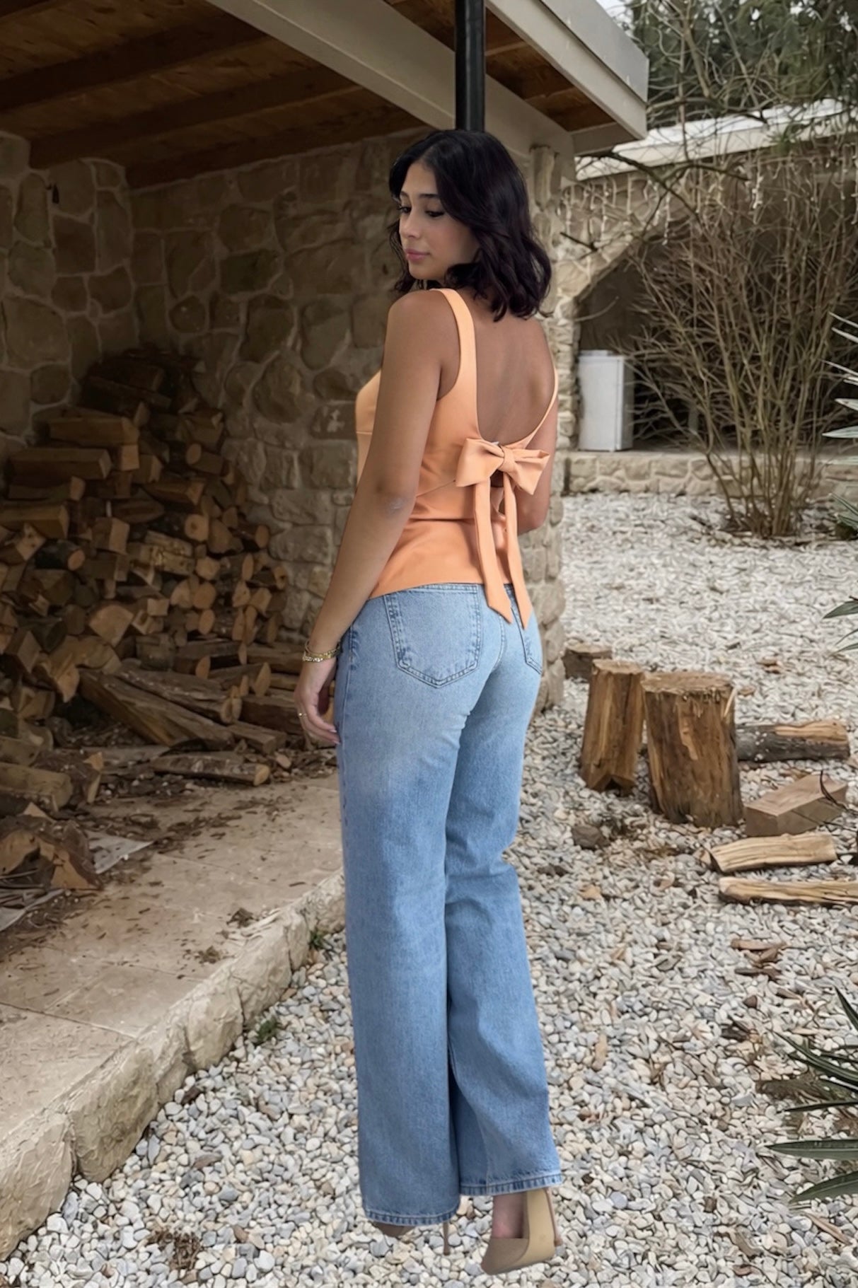 Woman in peach top and blue jeans standing outdoors near a stone wall and wooden logs.