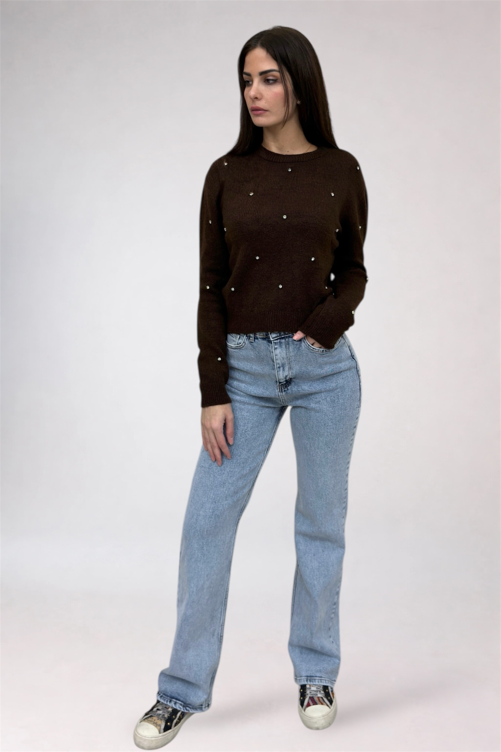 Woman wearing a brown sweater with pearl embellishments and blue jeans on a white background