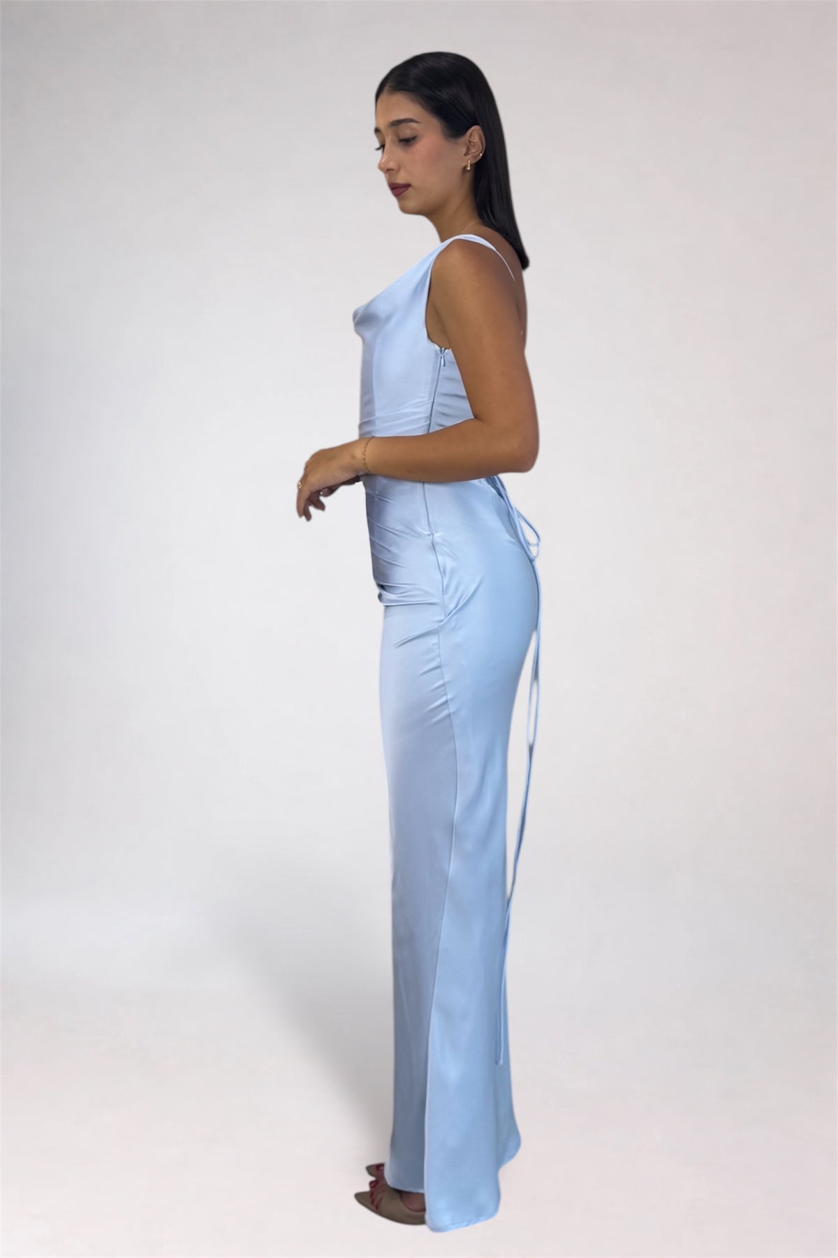 A woman modeling a light blue sleeveless maxi dress with a fitted bodice, side tie detail, and a long side slit, shown in profile against a plain light gray studio background.
