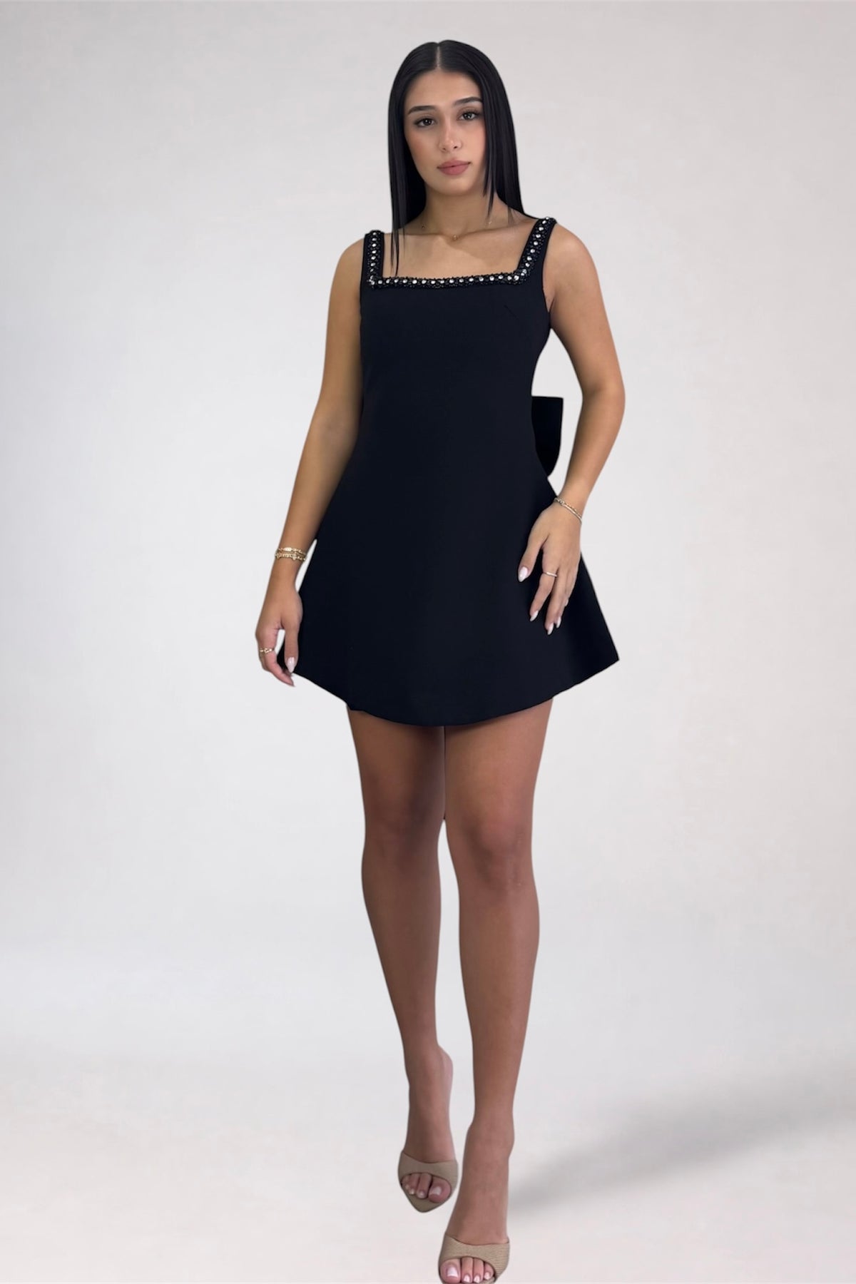 A woman modeling a black sleeveless mini dress with a square neckline and silver rhinestone trim, standing against a plain light gray studio background.