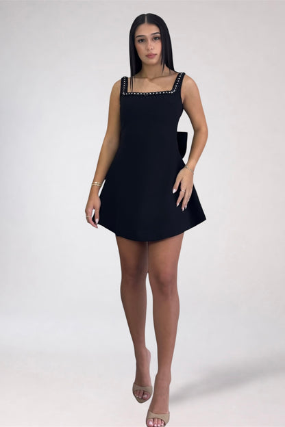 A woman modeling a black sleeveless mini dress with a square neckline and silver rhinestone trim, standing against a plain light gray studio background.