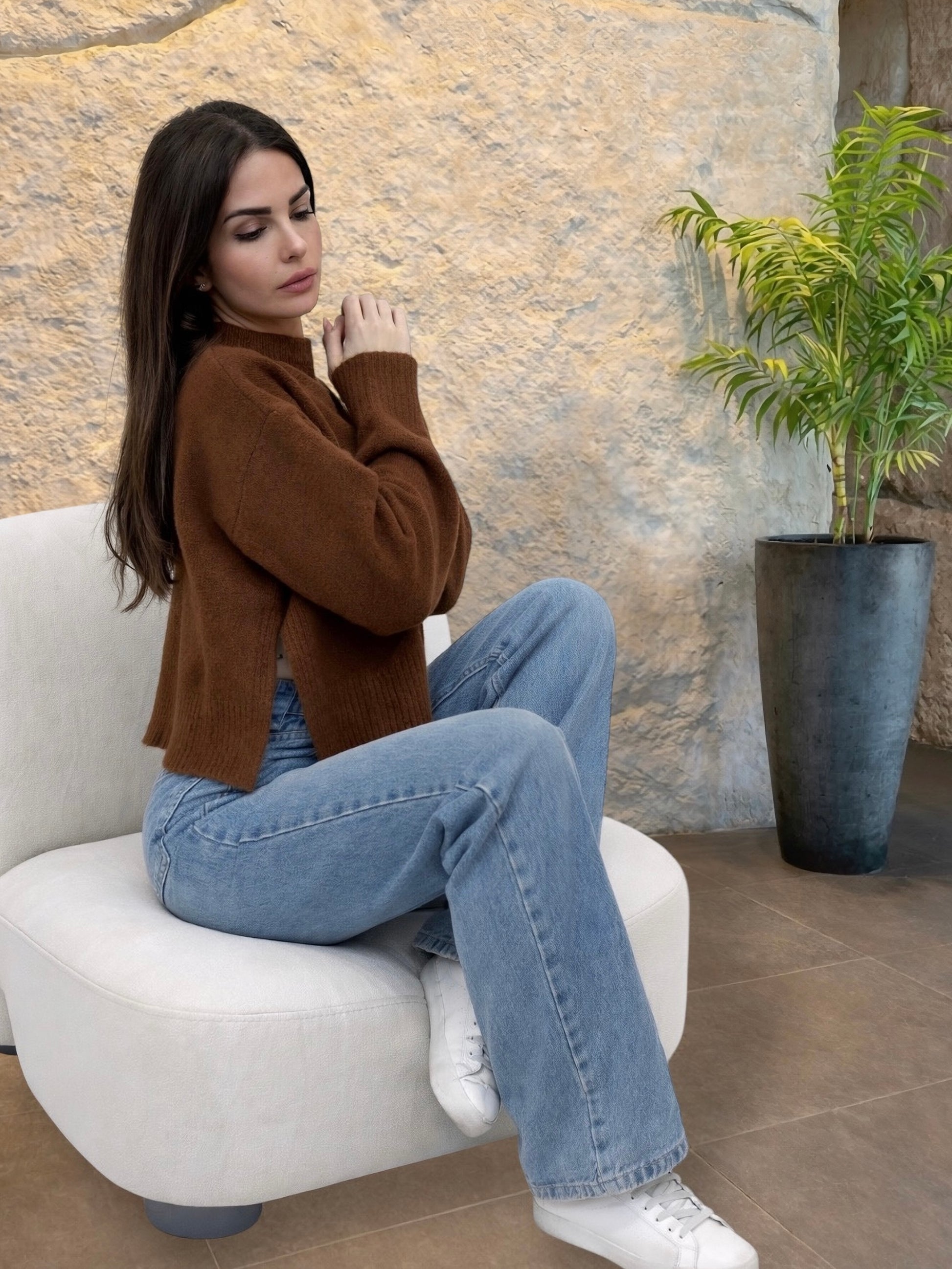 Woman sitting on a white chair wearing a brown sweater and blue jeans, with a stone wall and plant in the background.
