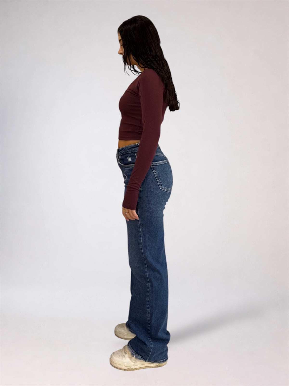 A woman shown in profile wearing a dark burgundy long-sleeve crop top, blue flare jeans, and off-white sneakers against a plain light gray studio background.