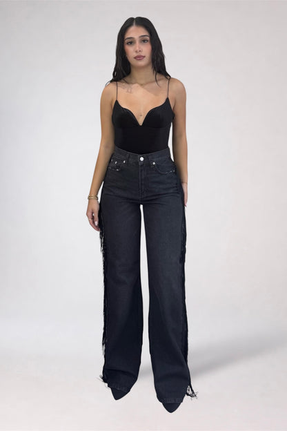 A woman modeling a black spaghetti-strap camisole top paired with high-waisted, wide-leg black denim jeans featuring fringe detailing along the outer seams, set against a plain light gray studio background.