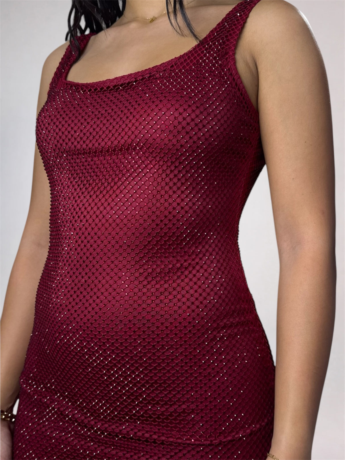 A close-up of a woman wearing a sleeveless scoop-neck dress covered in shimmering dark red sequins or mesh-like embellishment against a plain light gray background.