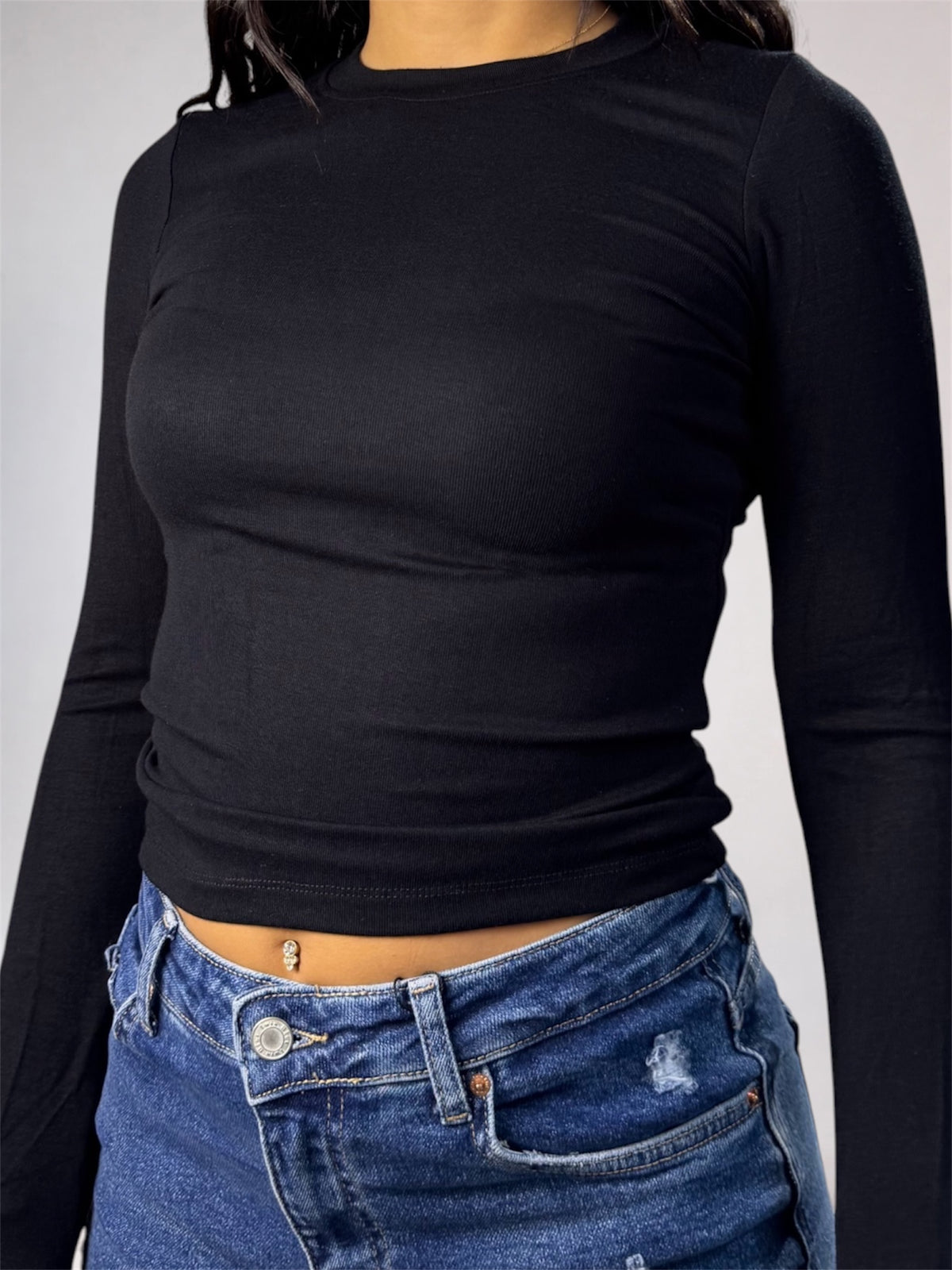 A person wearing a fitted black long-sleeve crop top with blue jeans, shown from the neck to the hips against a plain light gray studio background.