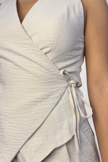 Close-up of a sleeveless wrap-style top in a light warm off-white or cream fabric, shown on a model with the side tie visible.