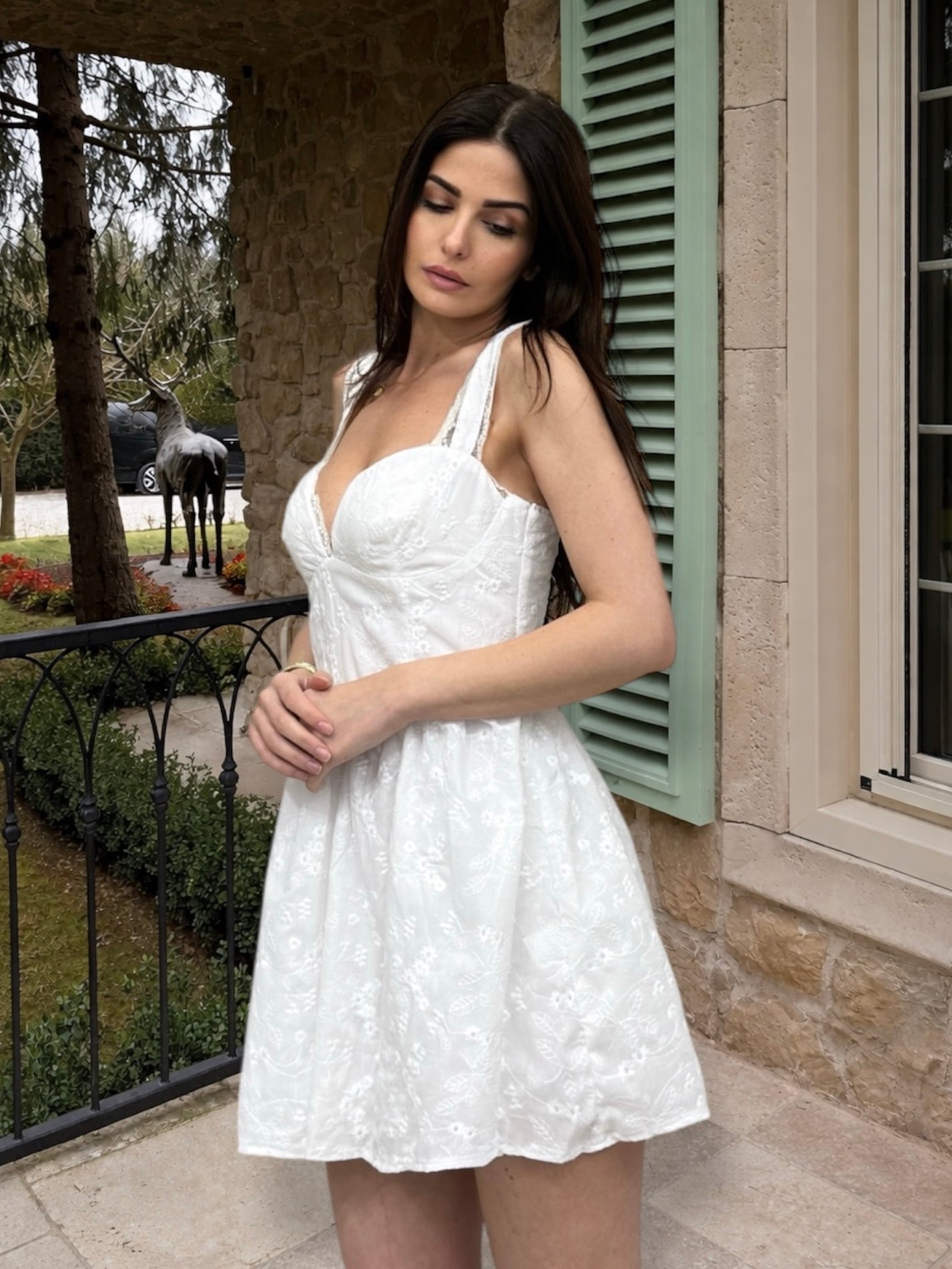 Woman in a white dress standing outdoors near a building with a horse in the background.