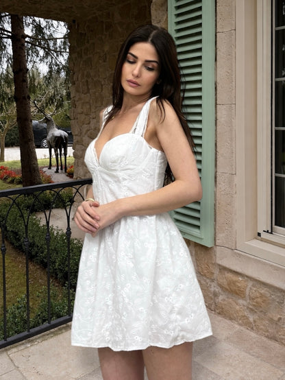 Woman in a white dress standing outdoors near a building with a horse in the background.