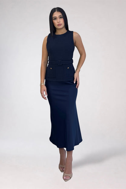 A woman modeling a sleeveless navy blue midi dress with a textured bodice, a matching belt with a decorative buckle, and a fitted skirt, set against a plain light gray studio background.