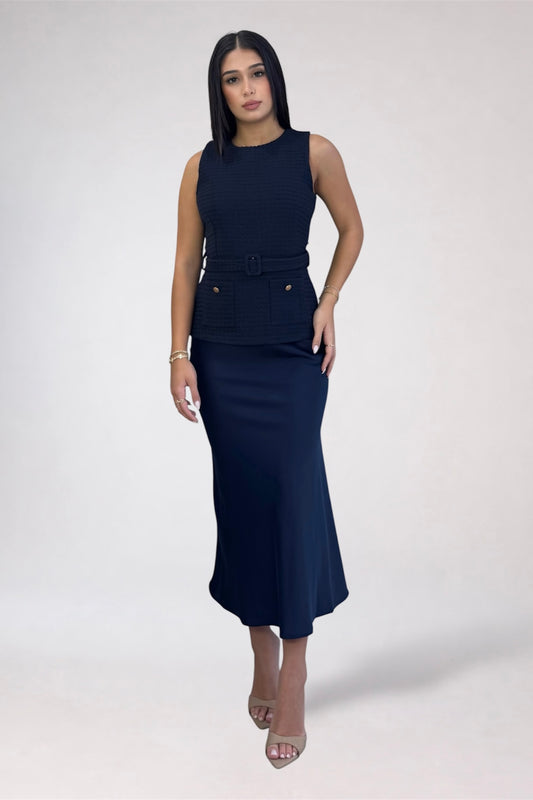 A woman modeling a sleeveless navy blue midi dress with a textured bodice, a matching belt with a decorative buckle, and a fitted skirt, set against a plain light gray studio background.