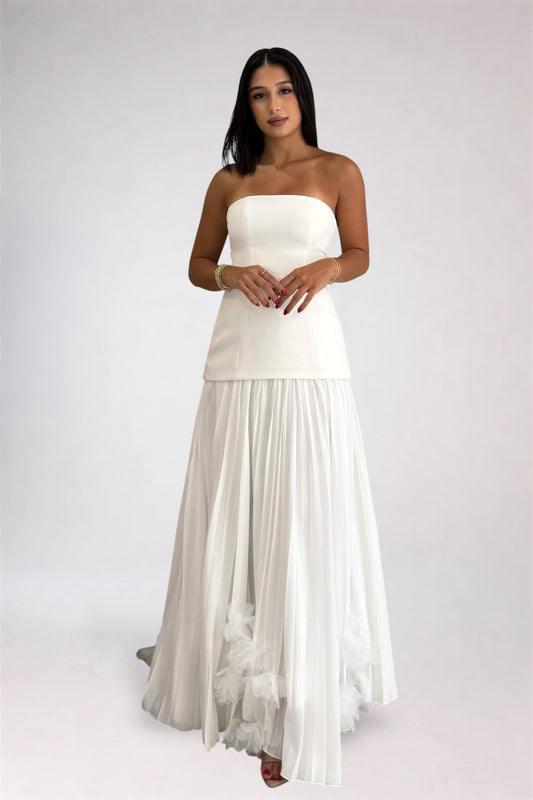 A woman modeling a strapless white maxi dress with a structured bodice and a flowing pleated skirt, photographed against a plain light gray studio background.