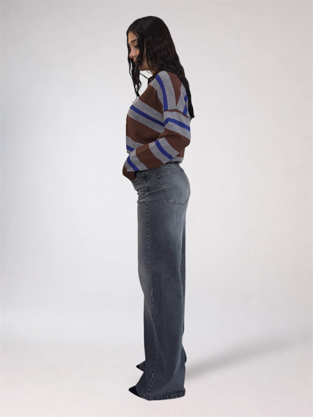 A woman stands in profile wearing a brown, blue, and gray striped knit sweater and a wide-leg gray denim skirt against a plain light gray studio background.