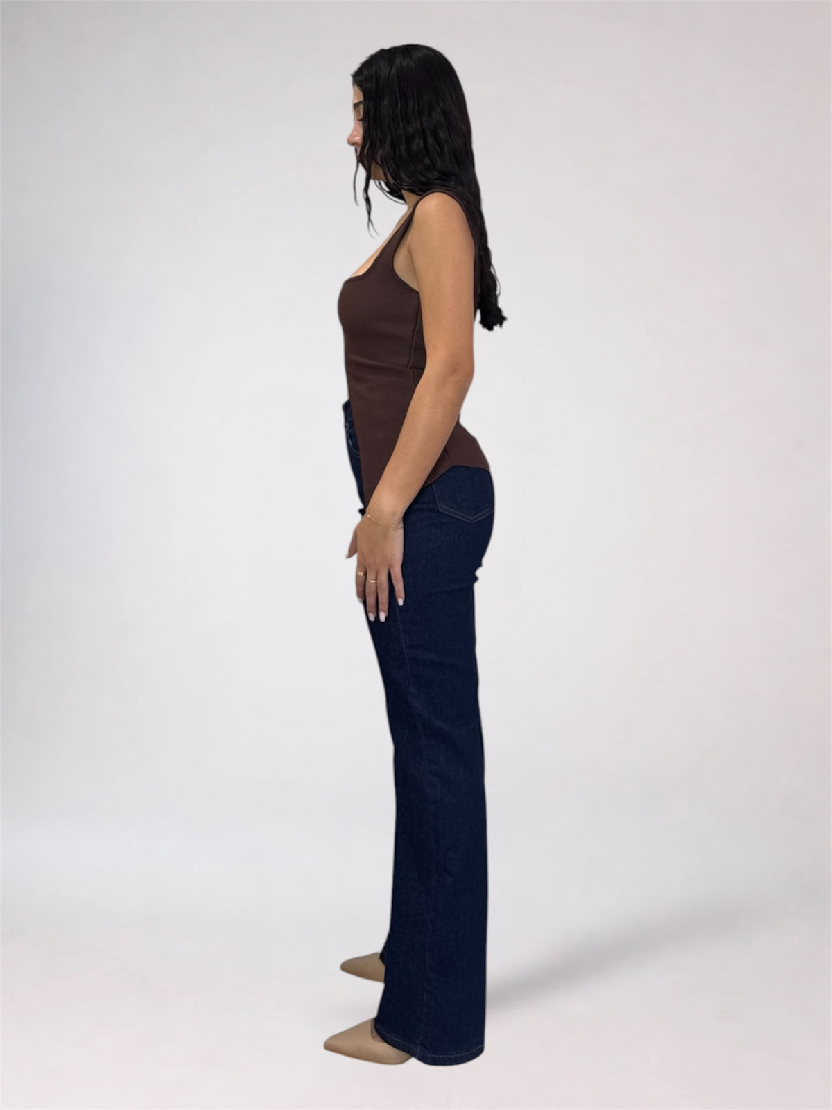 A woman shown in side profile wearing a brown ribbed tank top and dark blue flared jeans against a plain light gray studio background.