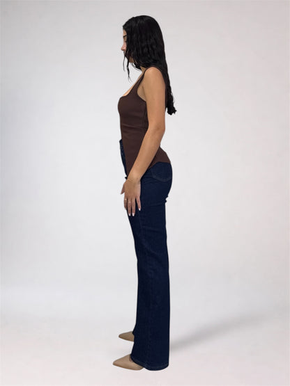 A woman shown in side profile wearing a brown ribbed tank top and dark blue flared jeans against a plain light gray studio background.