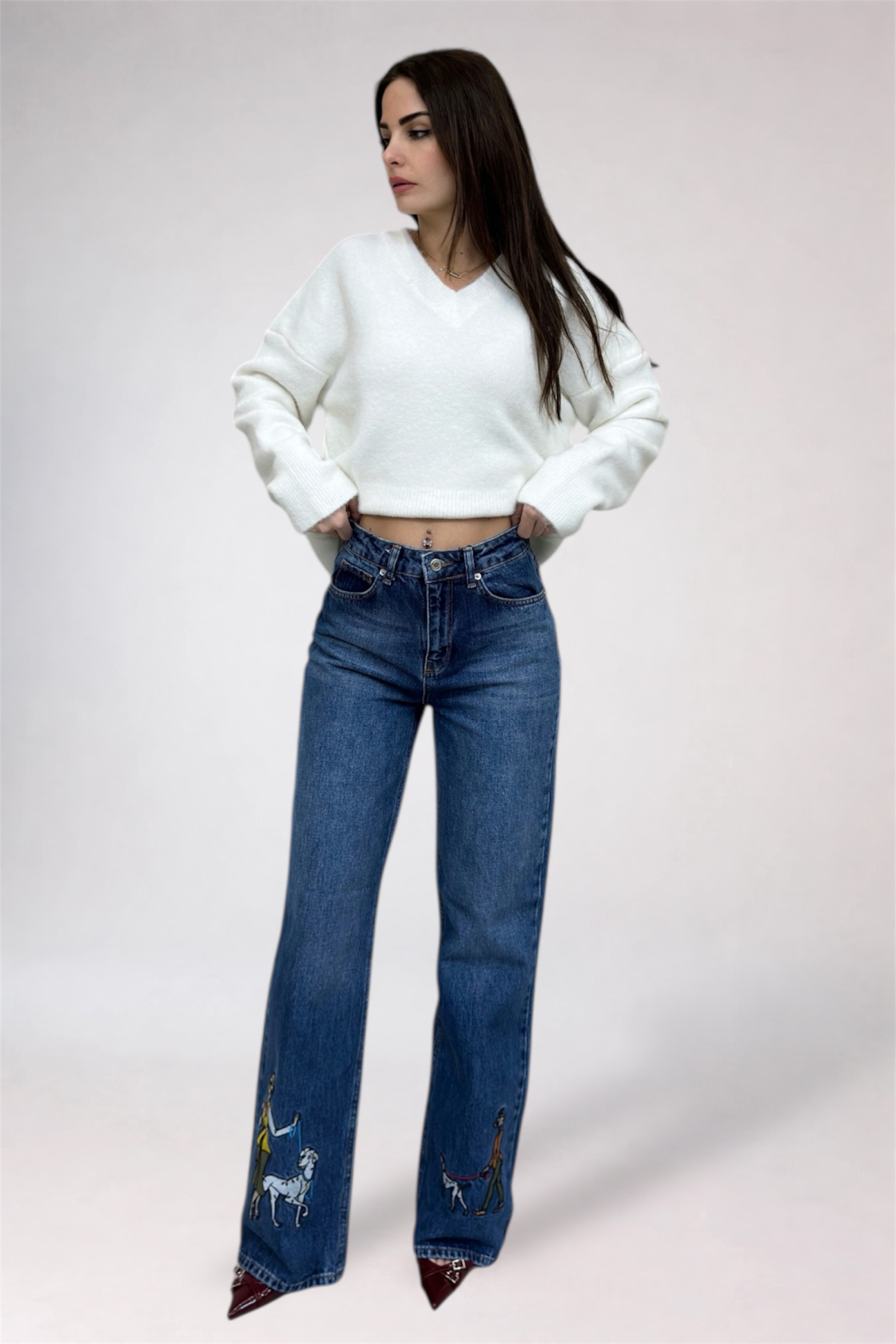 Woman wearing a white sweater and blue jeans with embroidery on a plain background