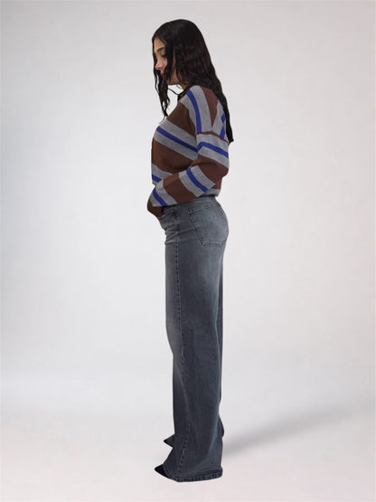 A woman stands in profile wearing a brown, blue, and gray striped knit sweater and a wide-leg gray denim skirt against a plain light gray studio background.