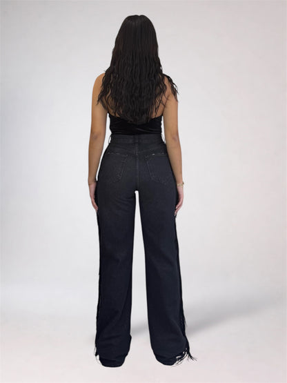 A woman shown from the back wearing a black strapless top and very dark wide-leg jeans with fringe detailing along the hems, set against a plain light gray studio background.