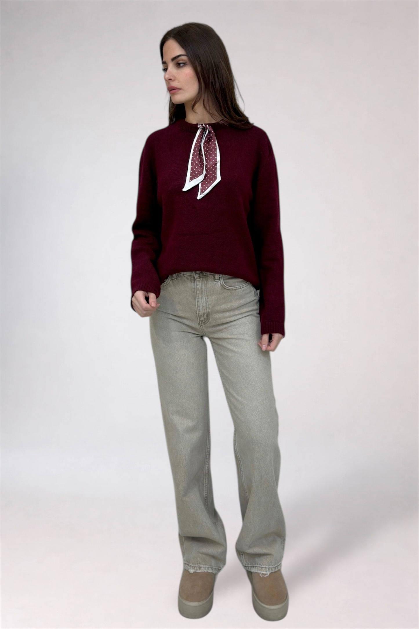 Woman wearing a burgundy sweater with a white scarf and light gray pants on a white background