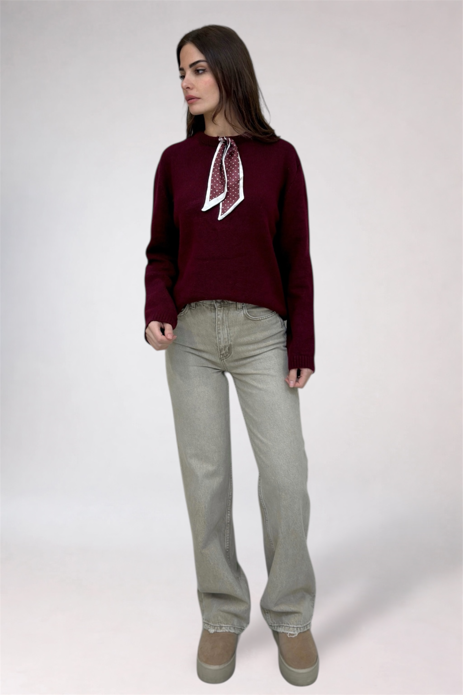 Woman wearing a burgundy sweater with a white scarf and light gray pants on a white background