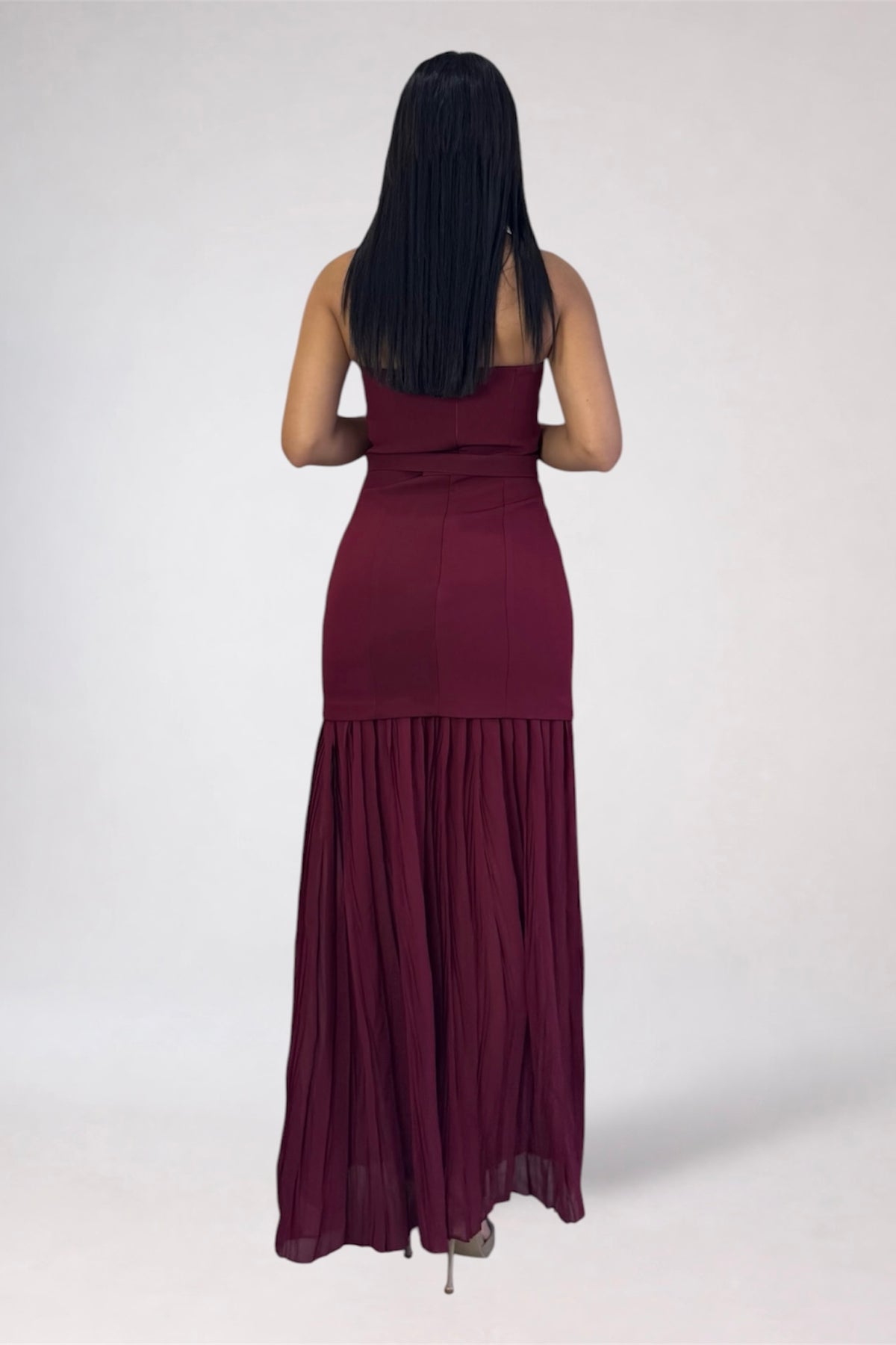 A woman viewed from behind wearing a strapless, floor-length burgundy maxi dress with a gathered waist and pleated skirt, standing against a plain light gray studio background.