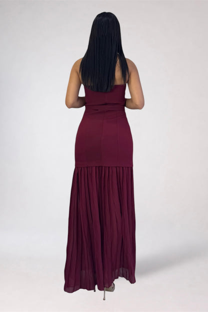 A woman viewed from behind wearing a strapless, floor-length burgundy maxi dress with a gathered waist and pleated skirt, standing against a plain light gray studio background.