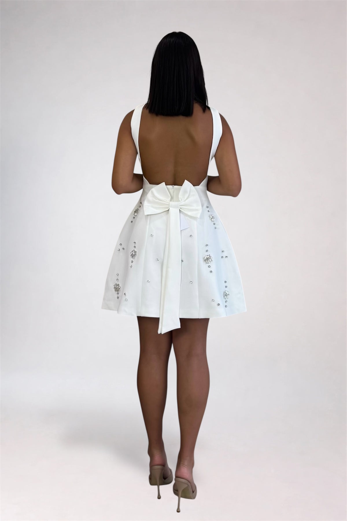 A woman viewed from behind wearing a white sleeveless mini dress with a large bow at the back waist and delicate floral embroidery, paired with nude high heels against a light gray studio background.