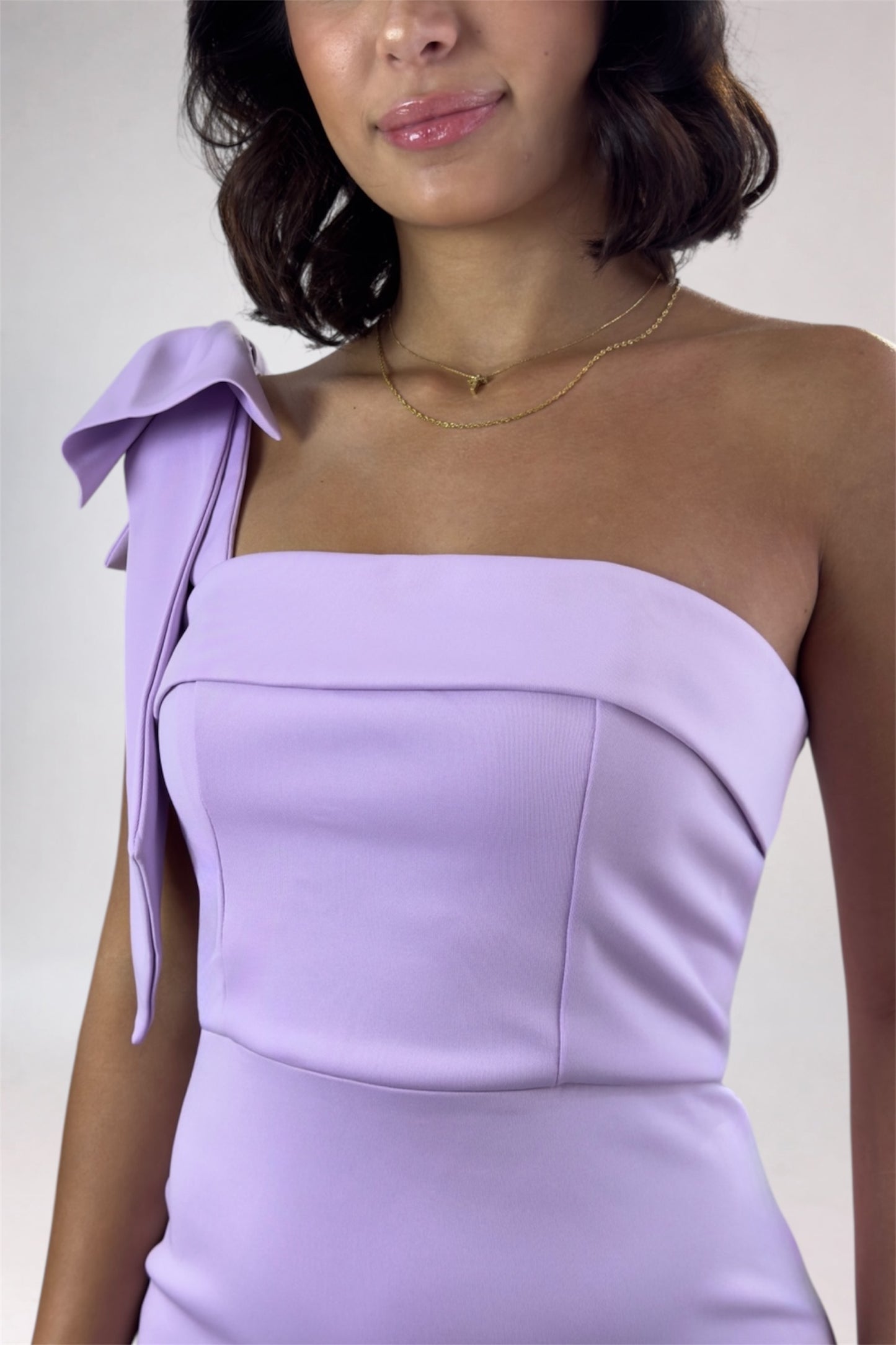 Woman wearing a lavender one-shoulder dress with a bow detail on a plain background