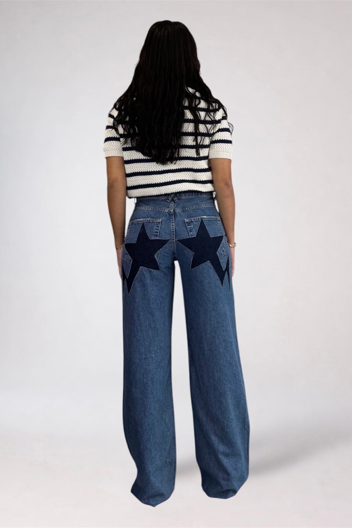 A woman viewed from behind wearing a black and white striped short-sleeve top and wide-leg blue jeans with large black star patches on the back pockets, against a plain light gray studio background.