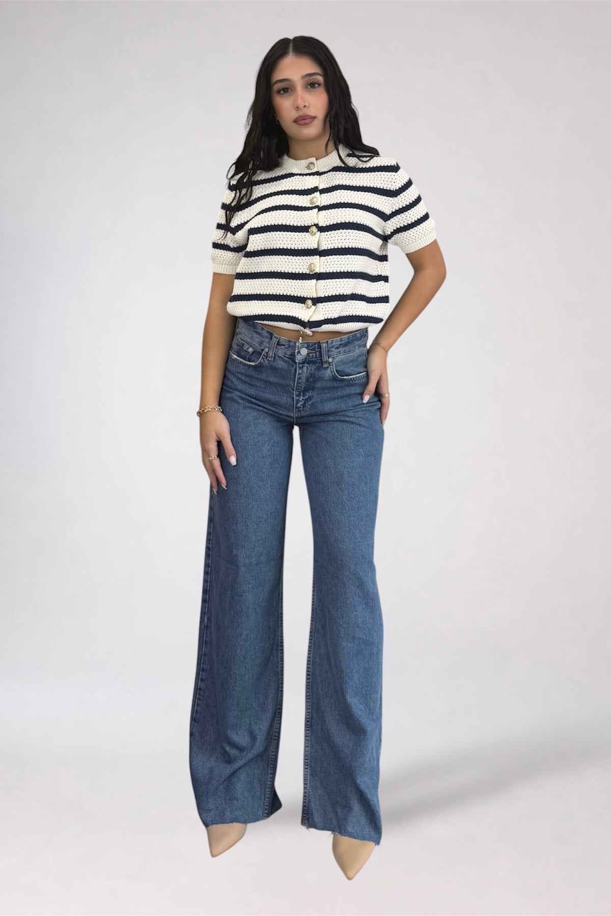 A woman modeling a short-sleeve knit top with navy and cream horizontal stripes, paired with high-waisted, wide-leg blue jeans, against a plain light gray studio background.