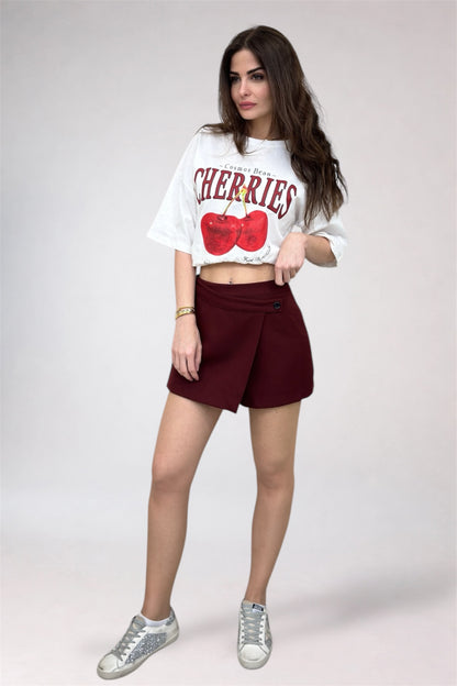 Woman wearing a white crop top with red cherries and maroon shorts on a white background