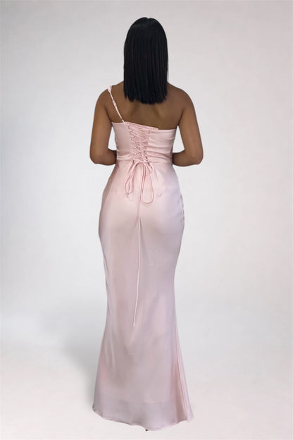 A woman viewed from behind wearing a light blush pink satin one-shoulder maxi dress with a lace-up back detail, set against a plain light gray studio background.