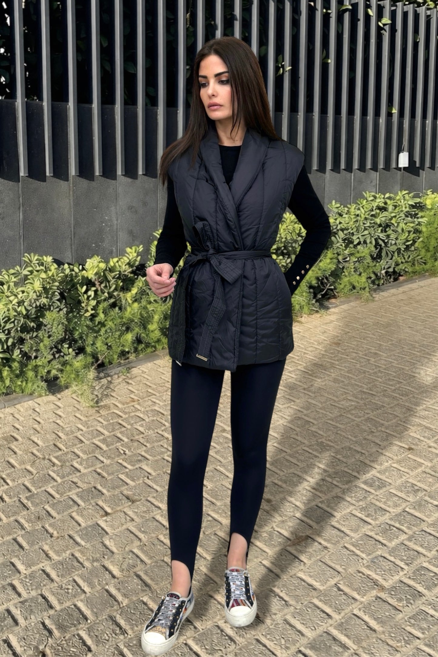 Woman wearing a black vest over a black top and leggings, standing on a paved path with greenery in the background.