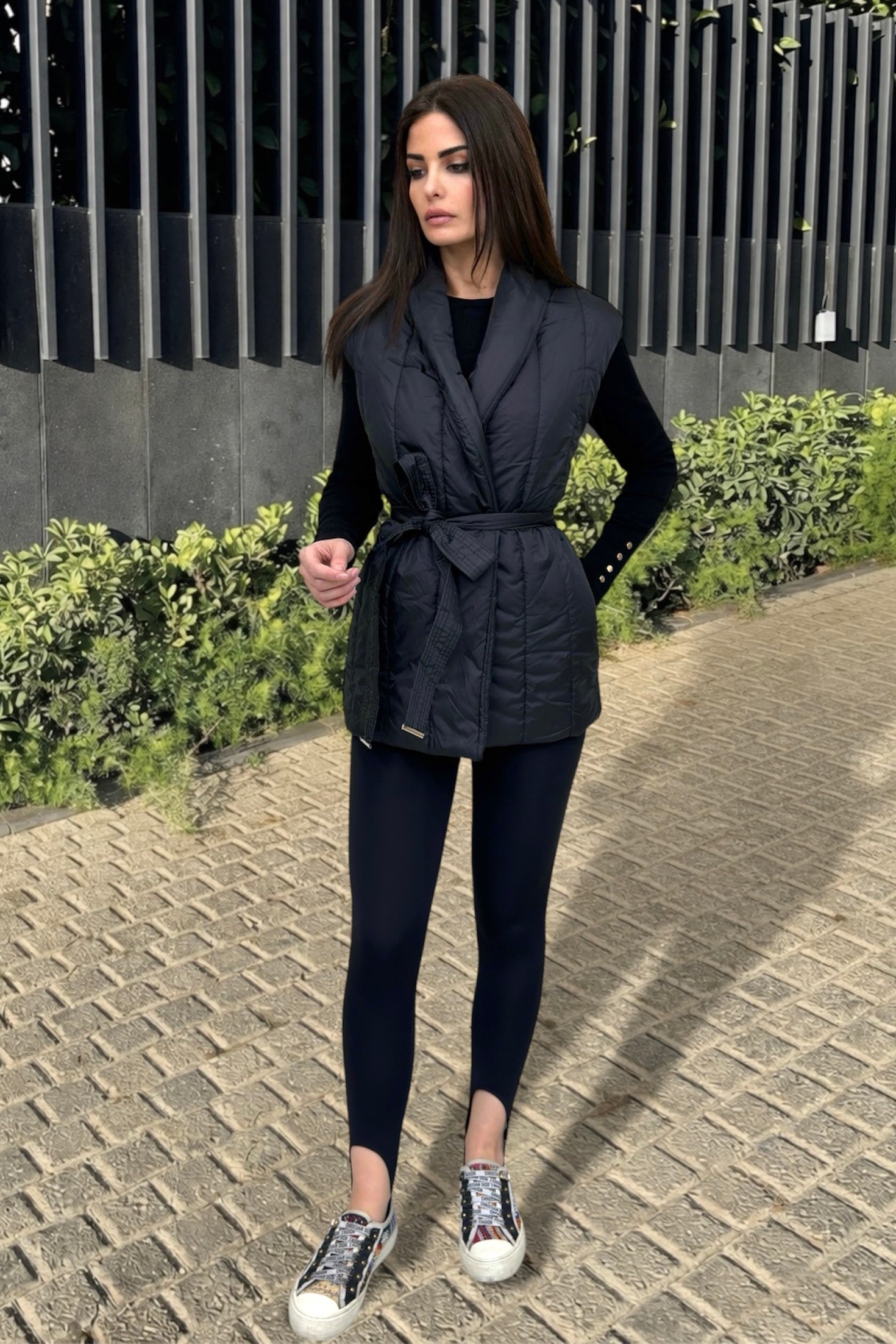 Woman wearing a black vest over a black top and leggings, standing on a paved path with greenery in the background.