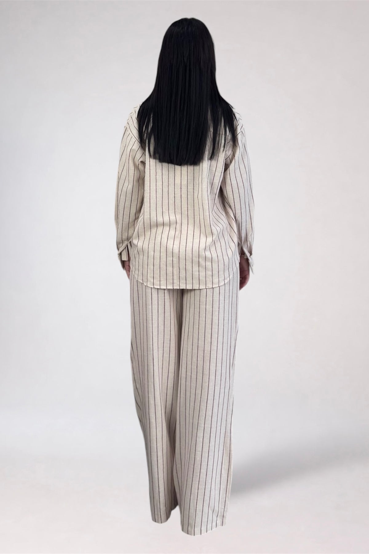 A woman viewed from behind wearing a matching two-piece pajama set with vertical stripes, consisting of a long-sleeve top and wide-leg pants, against a plain light gray background.