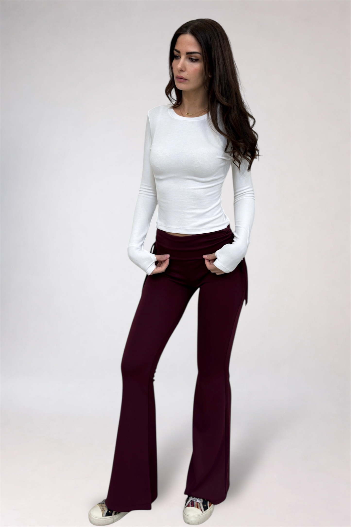 Woman wearing a white long-sleeve top and dark red flared pants on a plain background