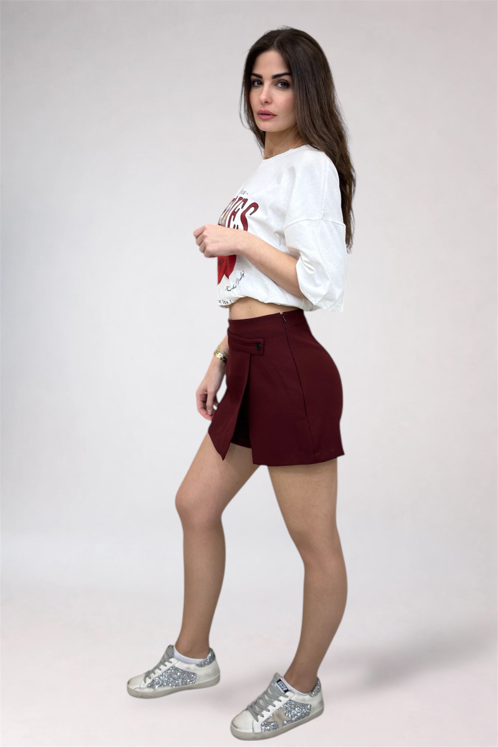Woman wearing a white crop top with red text, maroon skirt, and silver glittery sneakers on a light gray background