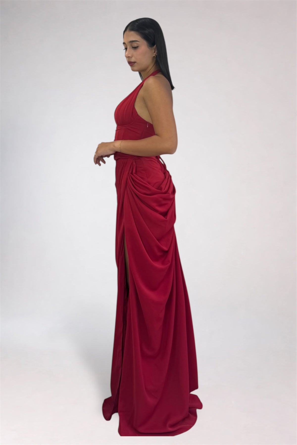 A woman modeling a floor-length, sleeveless red gown with a draped, gathered skirt and high side slit, photographed against a plain light gray studio background.