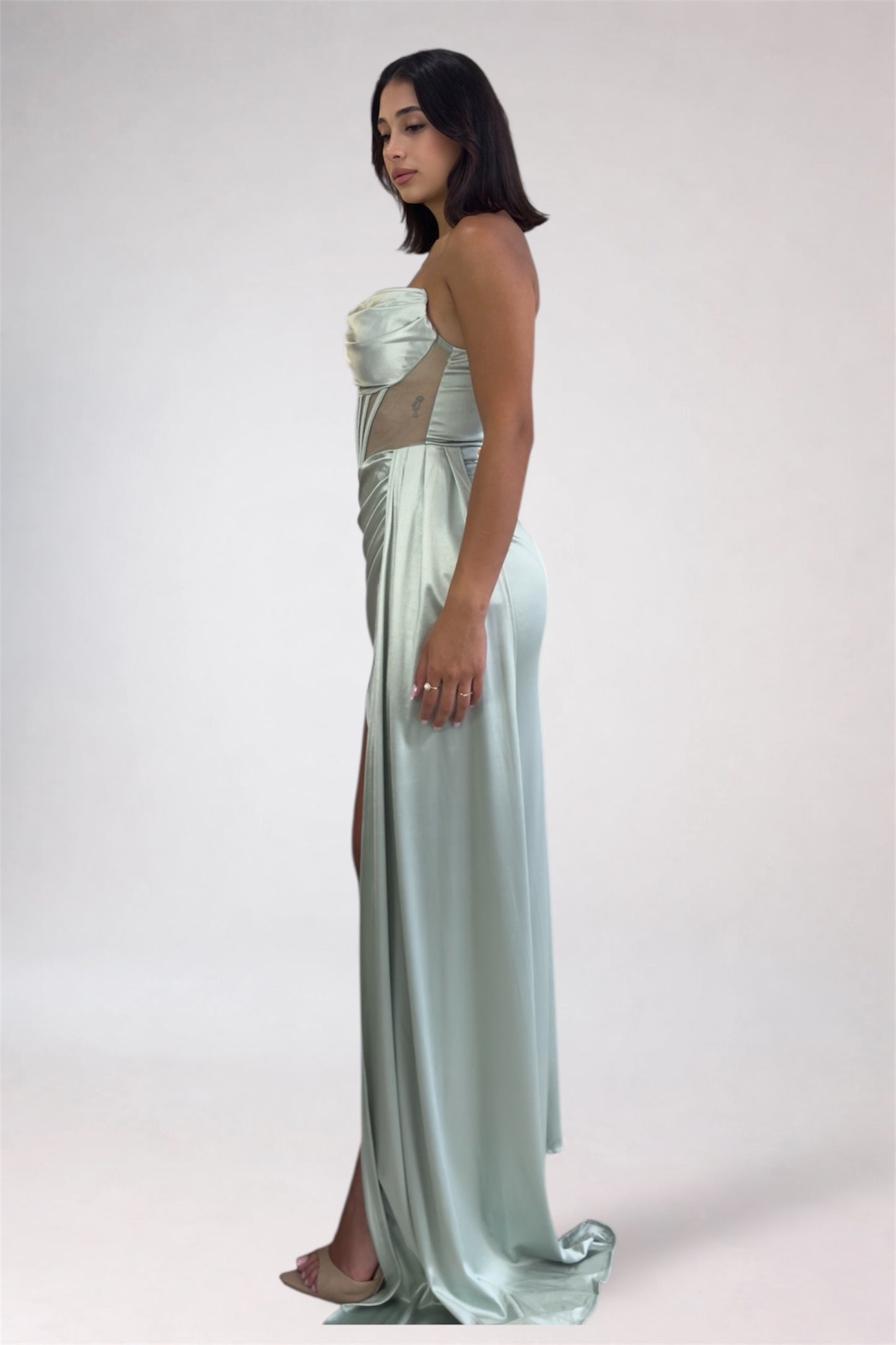 Woman wearing a strapless, light-colored evening gown with a slit on a plain background
