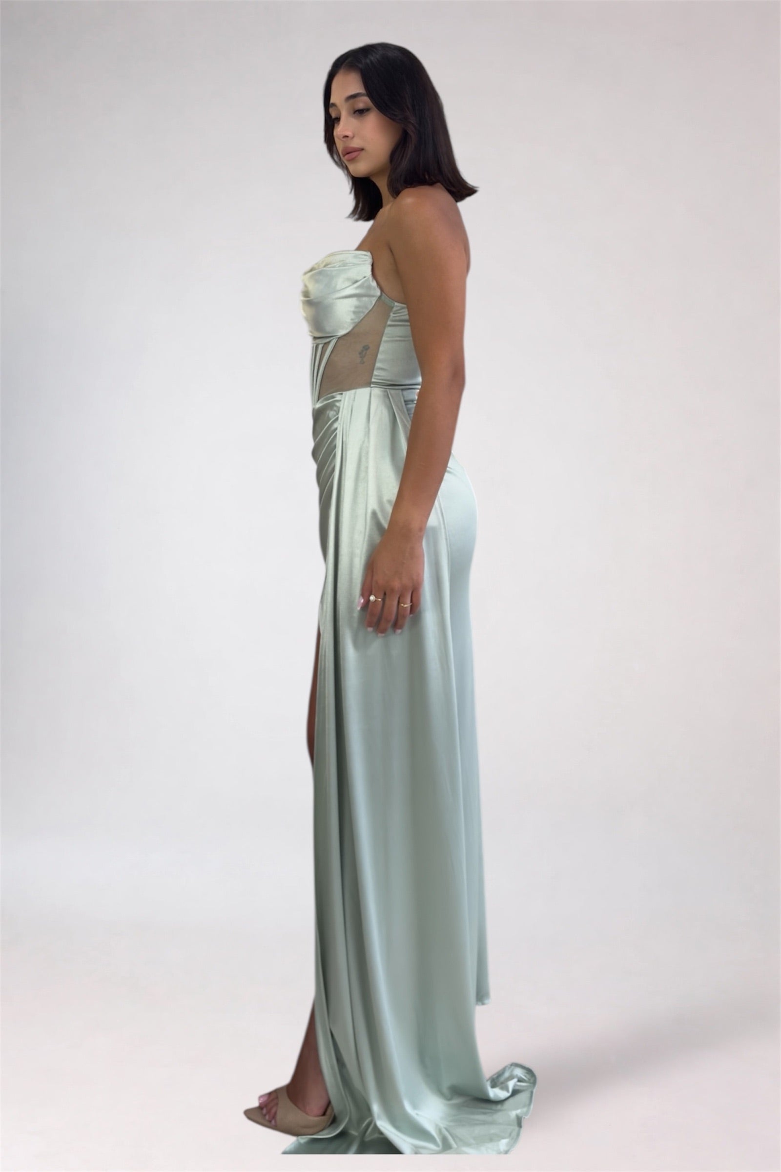 Woman wearing a strapless, light-colored evening gown with a slit on a plain background