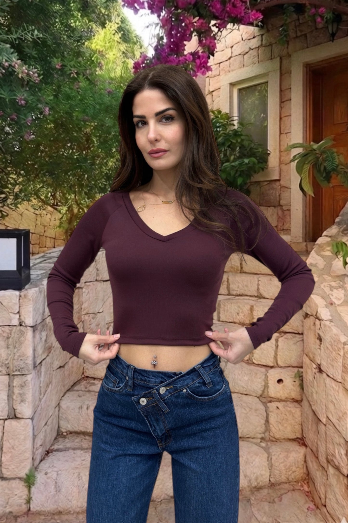Woman wearing a purple long-sleeve top and blue jeans standing outdoors with stone wall and plants in the background.