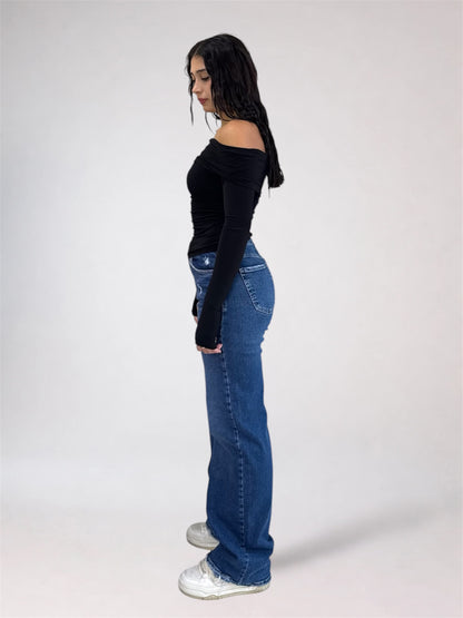 A woman standing in profile wearing a fitted black off-the-shoulder long-sleeve top, blue flare jeans, and white sneakers against a plain light gray studio background.