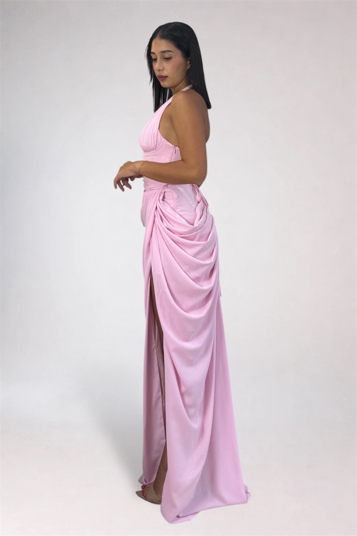 A woman modeling a light pink halter-neck maxi dress with a draped, gathered skirt and a high side slit, photographed against a plain light gray studio background.