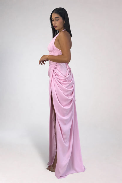 A woman modeling a light pink halter-neck maxi dress with a draped, gathered skirt and a high side slit, photographed against a plain light gray studio background.