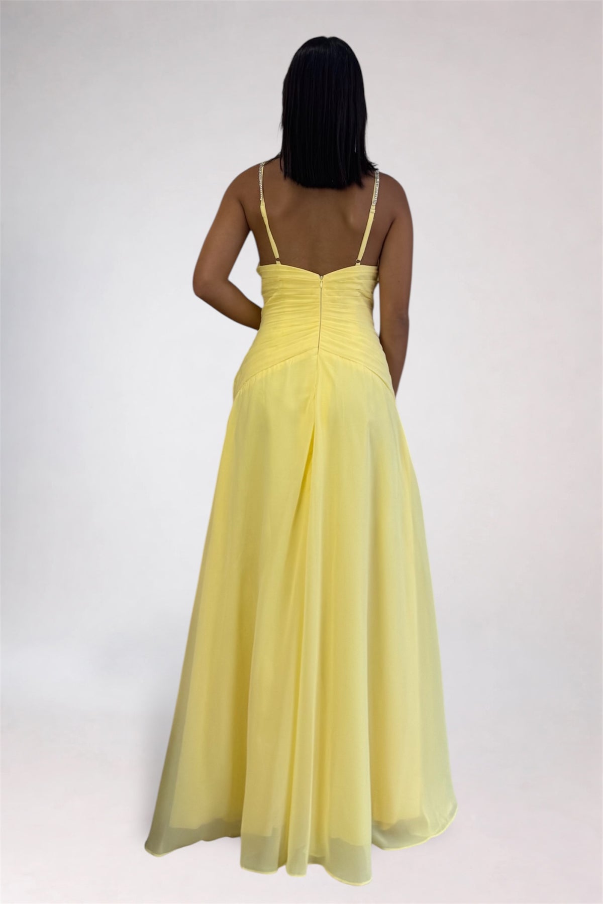 A woman viewed from the back wearing a pale yellow sleeveless maxi dress with thin straps, a fitted ruched bodice, and a flowing skirt against a plain light gray studio background.