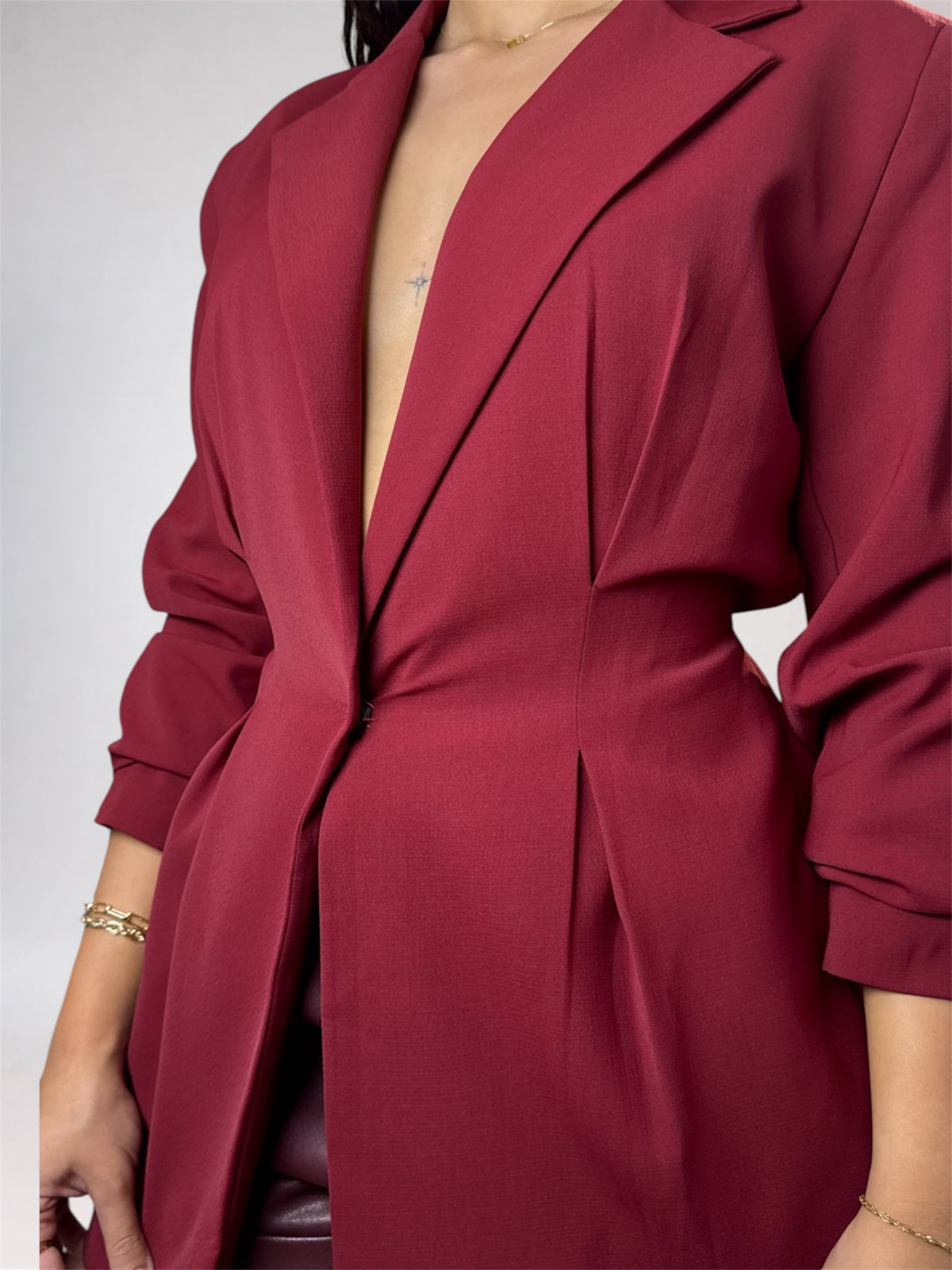 A close-up of a model wearing a deep burgundy longline blazer dress with a plunging V-neckline, structured shoulders, and long sleeves with buttoned cuffs.