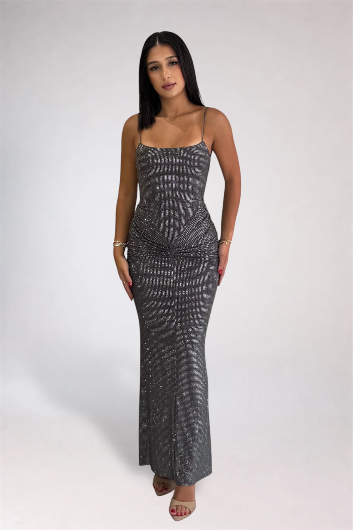 A woman modeling a floor-length, sparkly silver sequin gown with thin spaghetti straps and a gathered ruched bodice, standing against a light gray studio background.