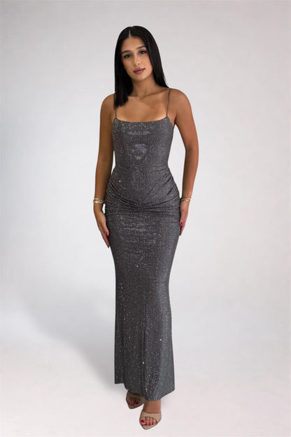 A woman modeling a floor-length, sparkly silver sequin gown with thin spaghetti straps and a gathered ruched bodice, standing against a light gray studio background.